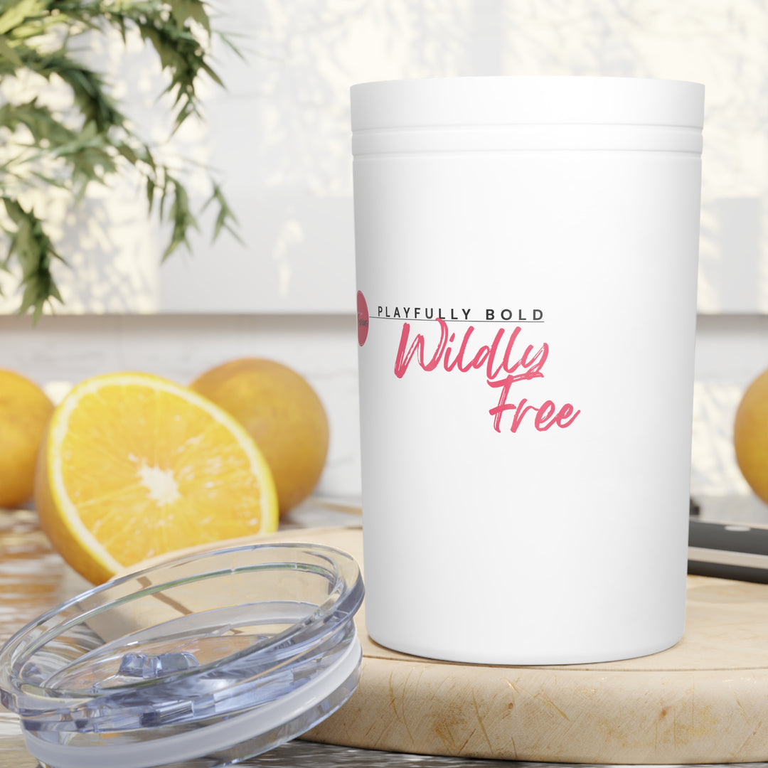 White tumbler with 'Playfully Bold, Wildly Free' text on a wooden surface with lemons and a plant in the background.