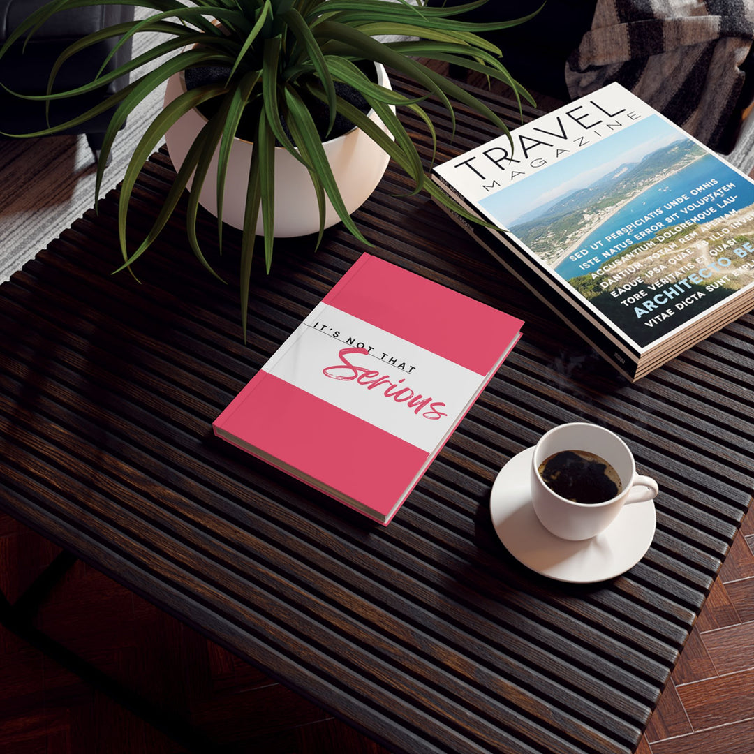 A hard backed journal with vibrant pink and white print on the cover, titled 'It's Not That Serious', placed on a table with a cup of coffee and a plant in the background, alongside a travel book.