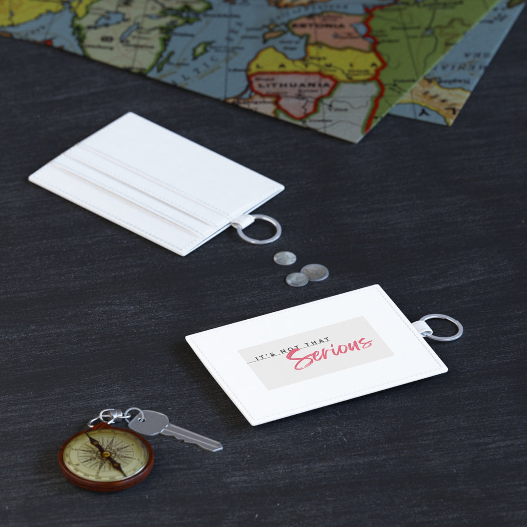 A white faux leather card holder with a red text design on one side, accompanied by a metal keyring, placed on a dark surface with a map and a compass.