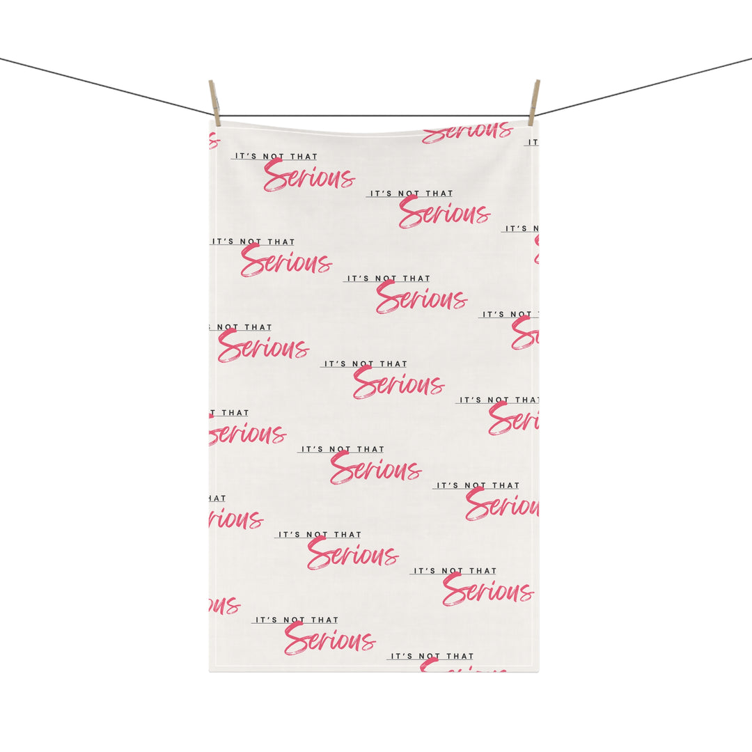 A white cotton tea towel with red text pattern hanging on a white shelf, with kitchen items and a plant in the background.