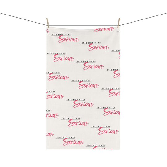 A white cotton tea towel with red text pattern hanging on a white shelf, with kitchen items and a plant in the background.