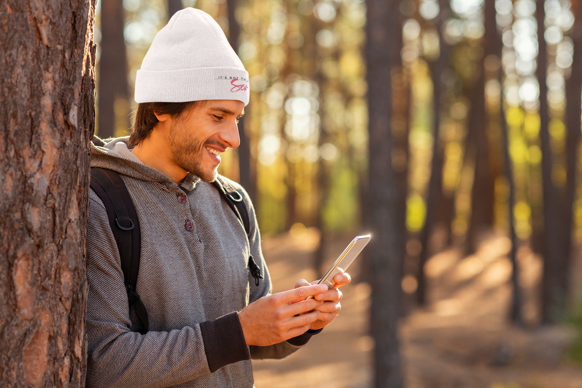 A person wearing a gray knit beanie with embroidery, standing in a forest setting and smiling while using a smartphone.