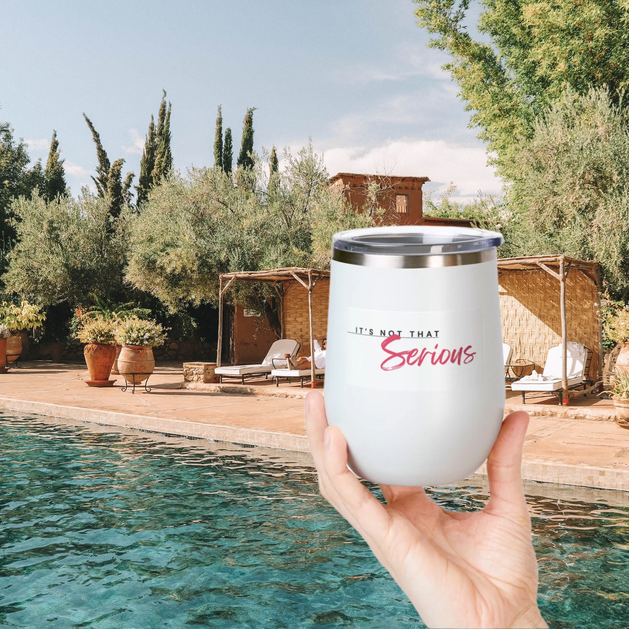 A hand holding a white 12oz wine tumbler with a printed phrase 'It's Not That Serious' on the side, in front of a pool and outdoor setting.