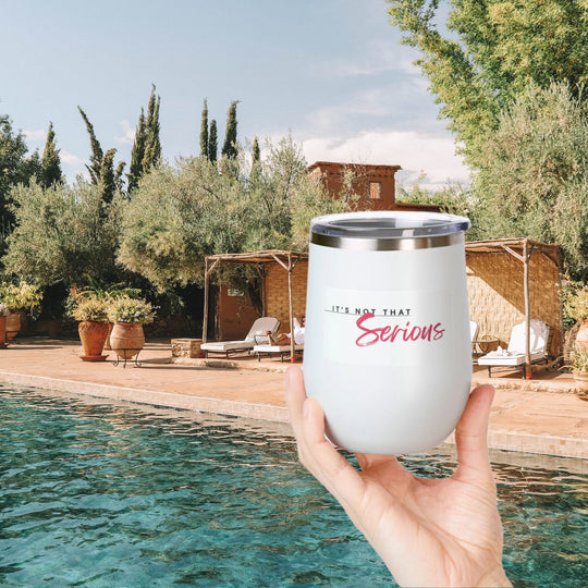 A hand holding a white 12oz wine tumbler with a printed phrase 'It's Not That Serious' on the side, in front of a pool and outdoor setting.