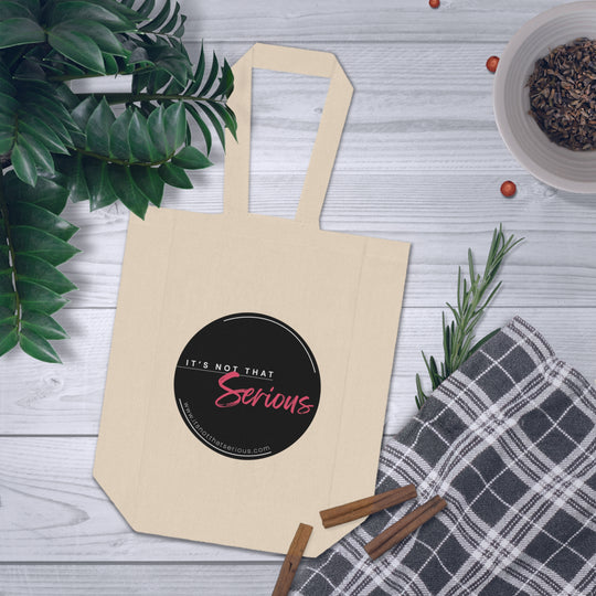 Beige tote bag with a black circular design on a wooden surface with plants and cinnamon sticks.