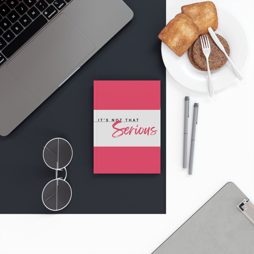 A hard backed journal with vibrant pink and white print on the cover, titled 'It's Not That Serious', placed on a desk with a laptop