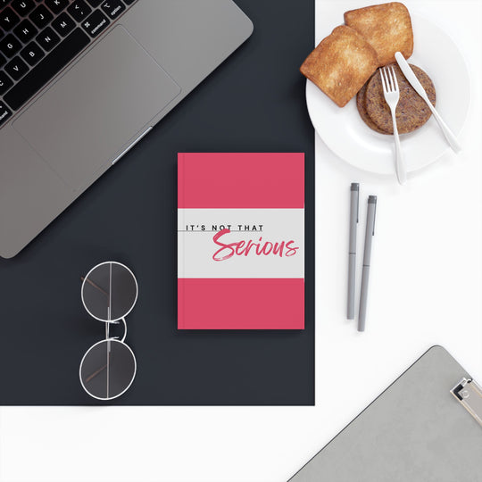 A hard backed journal with vibrant pink and white print on the cover, titled 'It's Not That Serious', placed on a desk with a laptop