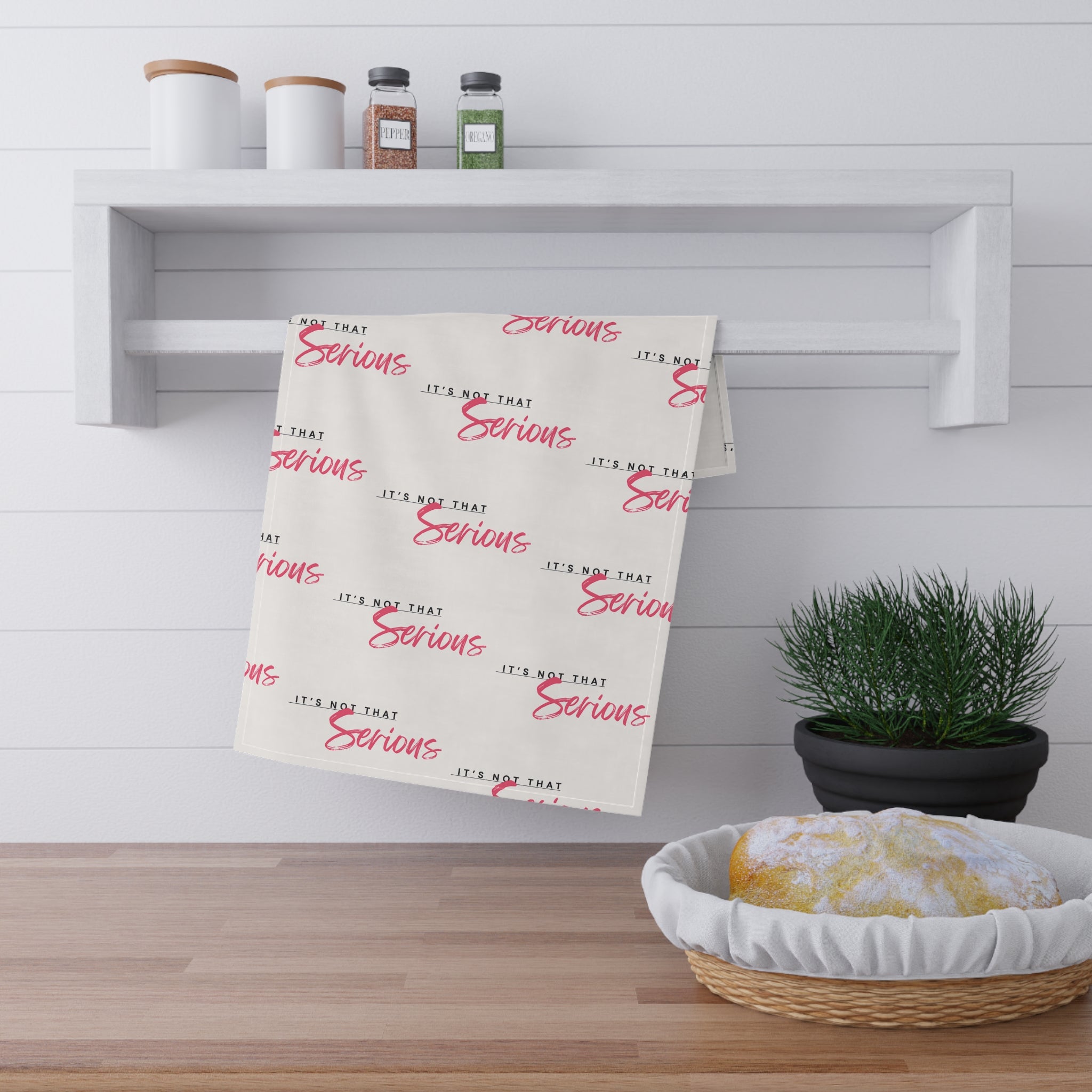 A white cotton tea towel with red text pattern hanging on a white shelf, with kitchen items and a plant in the background.