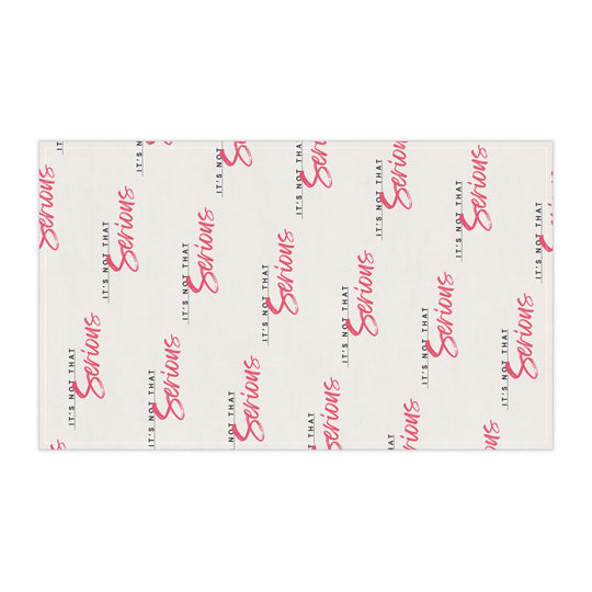 A white cotton tea towel with red text pattern hanging on a white shelf, with kitchen items and a plant in the background.