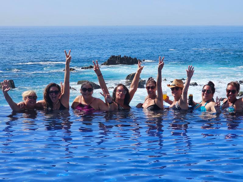 A photo of eight middle aged women in a pool overlooking the ocean with their arms in the air