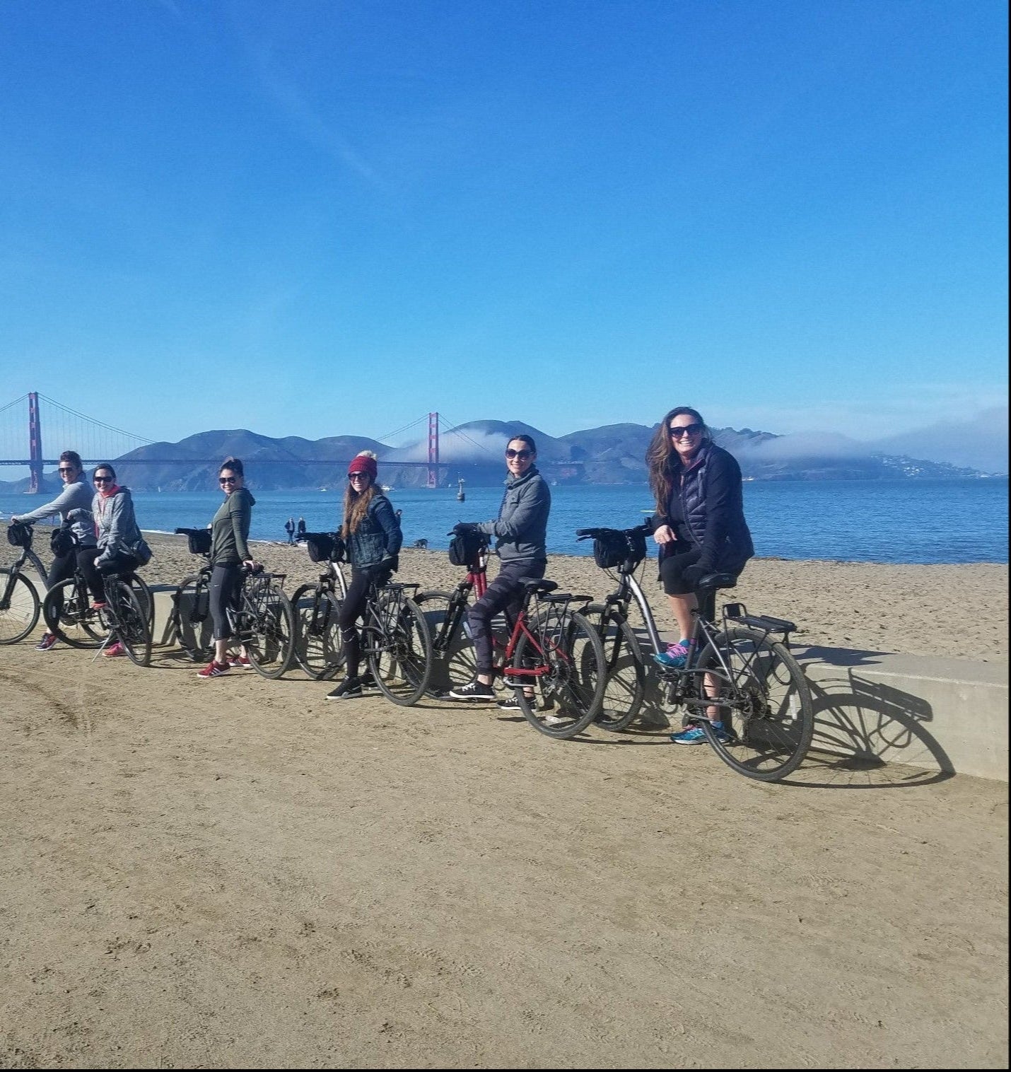 Six women on bicycles in a line in front of the Golden Gate bridge