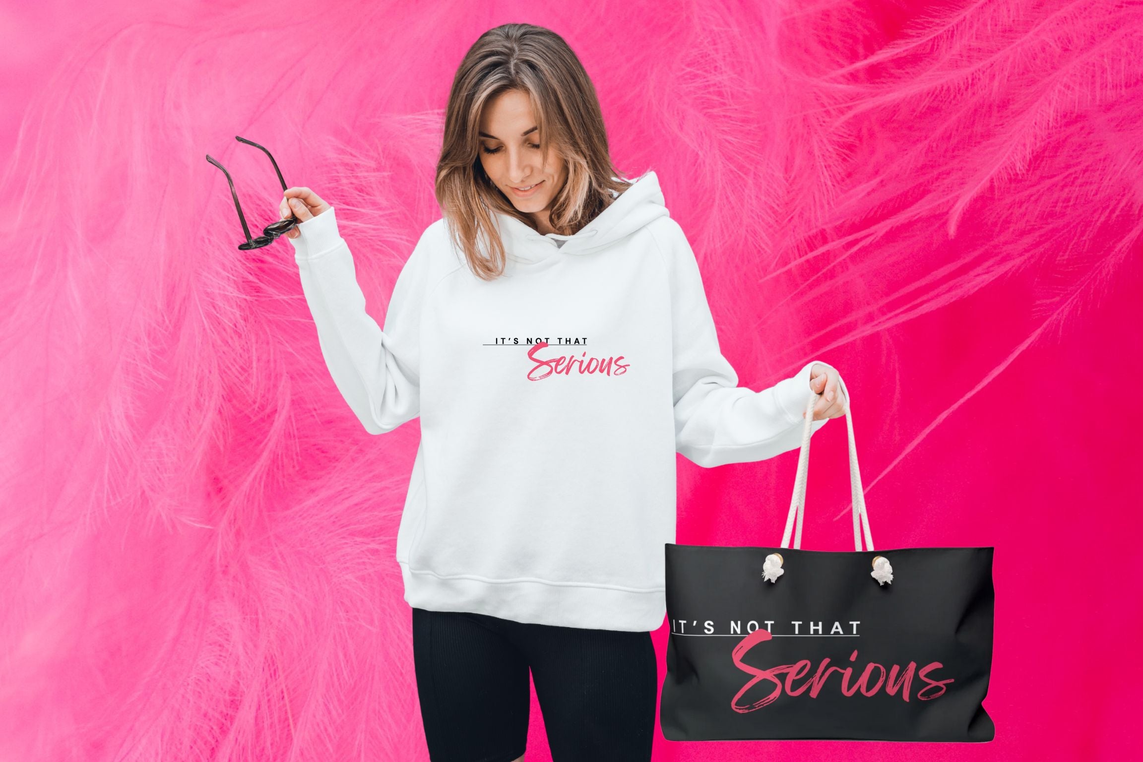Women in a white It's Not That Serious logo hoodie with a branded weekender bag with a hot pink feather background