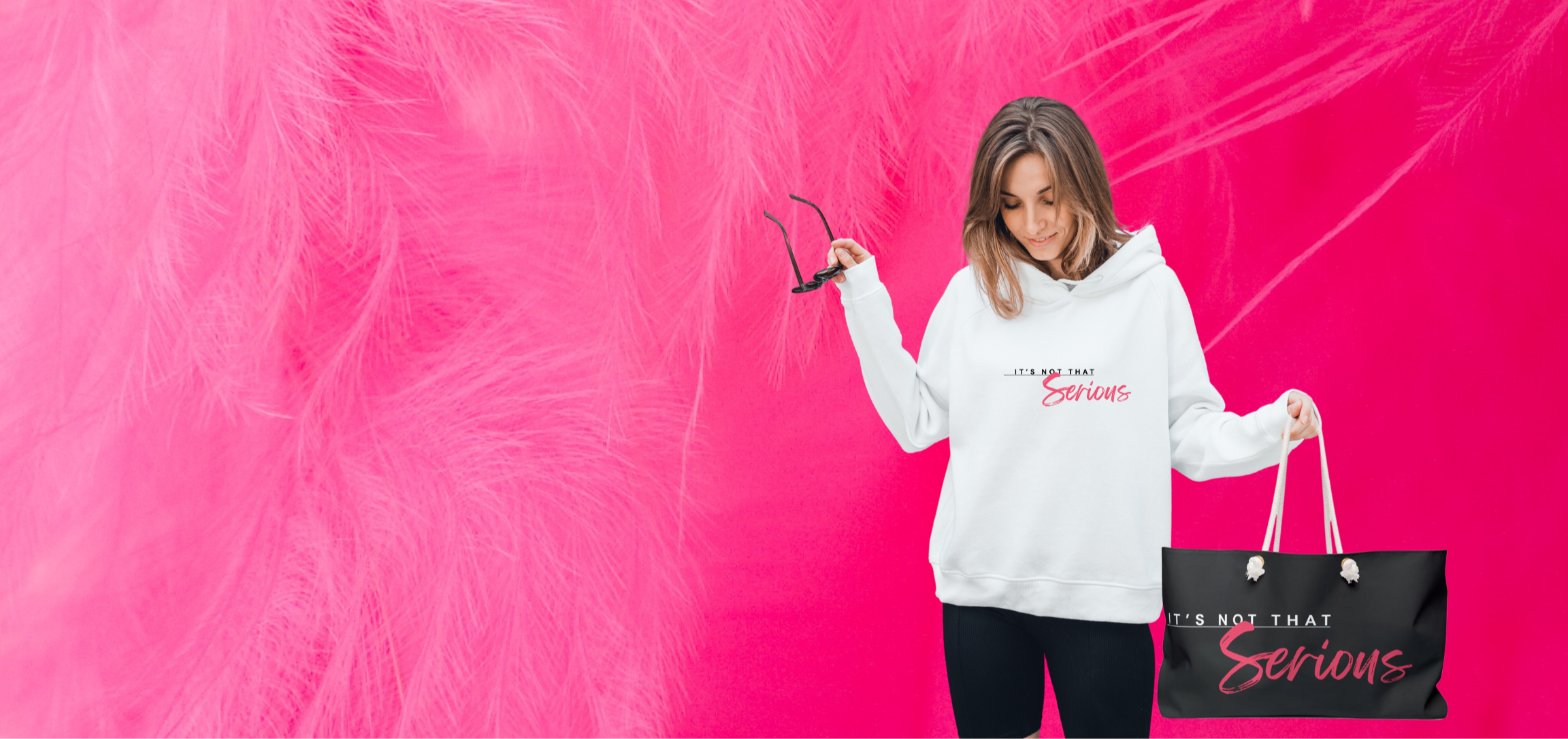 Women in a white It's Not That Serious logo hoodie with a branded weekender bag with a hot pink feather background
